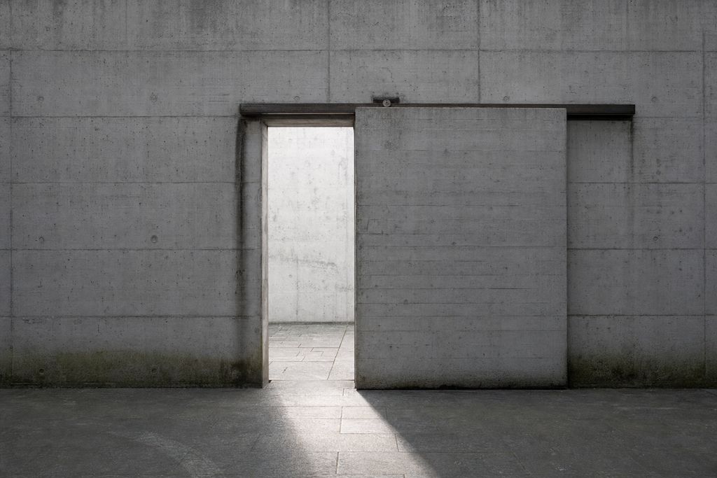 A long blank concrete wall with one heavy sliding panel drawn open, letting a band of daylight across the paving.