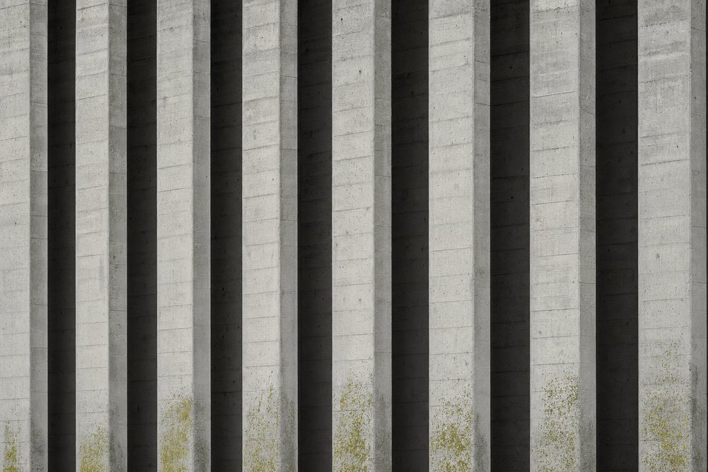 A colonnade of identical narrow concrete piers receding across the frame, each separated by a slot of shadow.